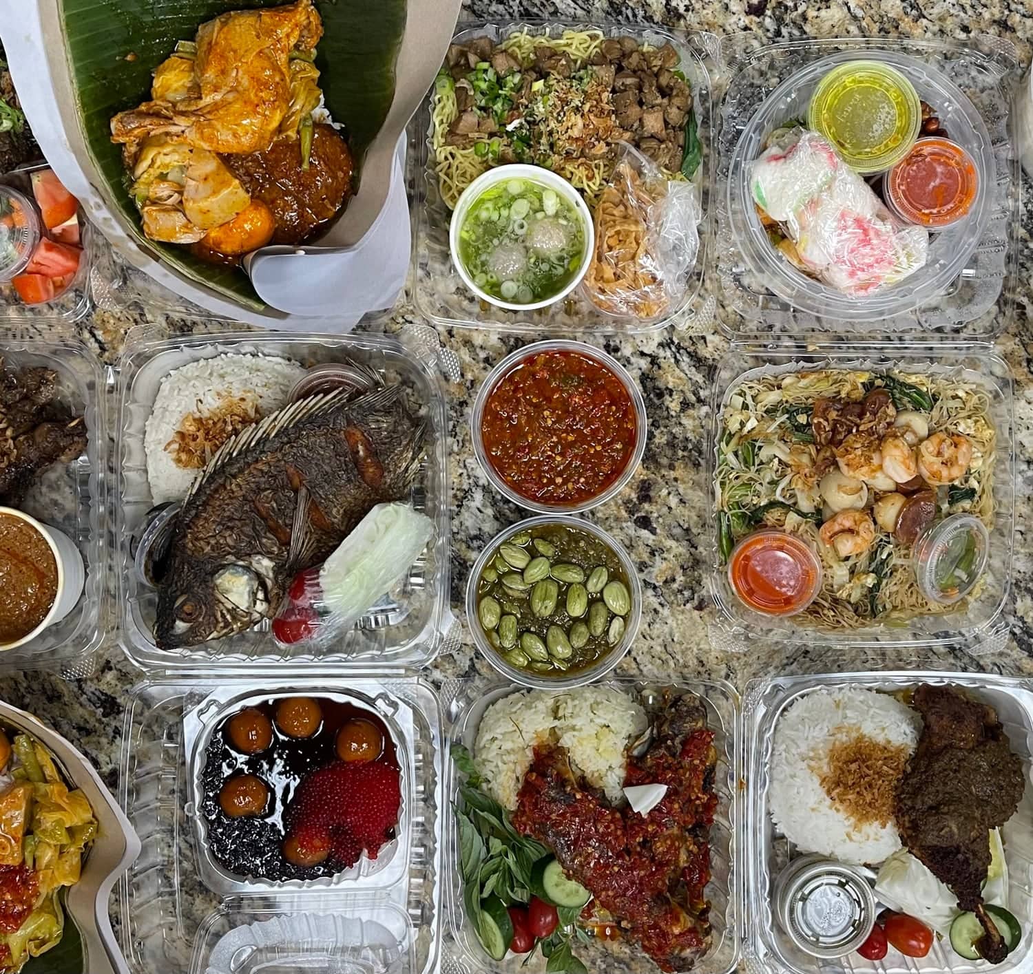 The counter before opening, shot from above: packed takeout containers filling the frame — a whole fried fish, sambals, petai, noodles with prawns, a black sticky rice sweet, and nasi padang on banana leaf
