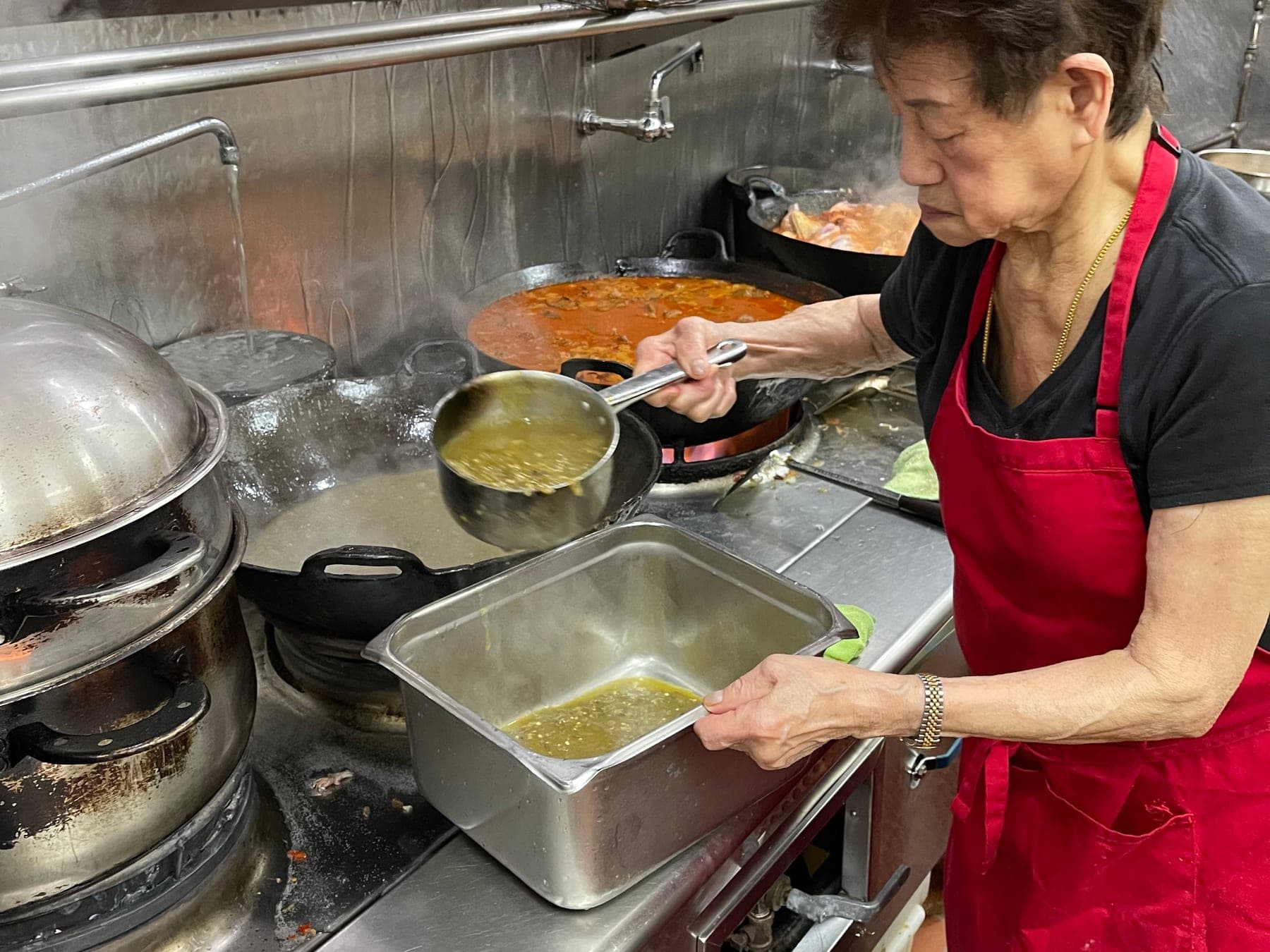 Siu Chen in a red apron, ladling sambal hijau from a wok into a steel pan, steam rising around her